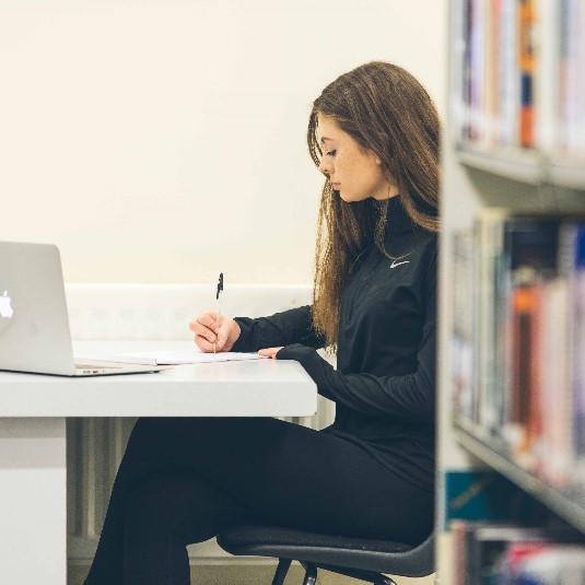 Library user seated at desk writing, laptop and bookshelves in foreground